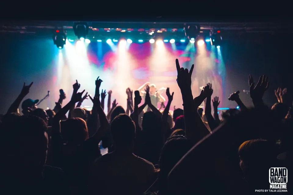 Silhouetted crowd with raised hands and rock horn gestures facing a stage lit in blue, pink, and white lights.