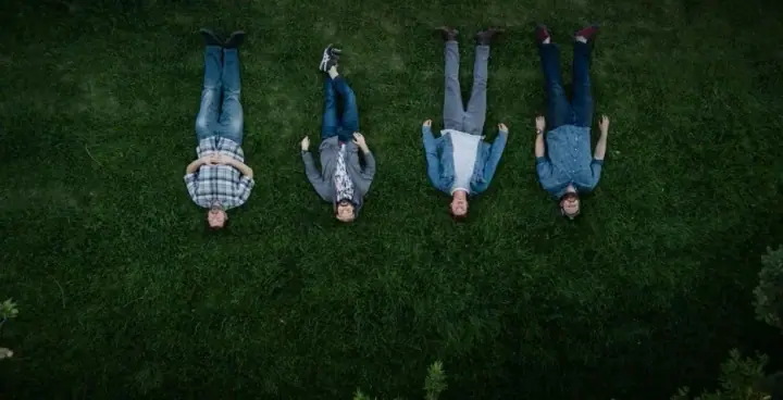 Four people in jeans and casual clothes lie on their backs in green grass, photographed from above.