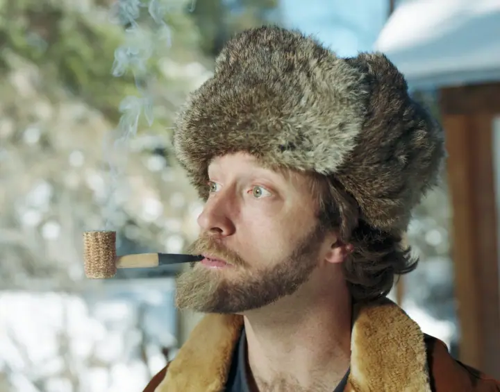 A bearded man in a fur hat and shearling collar holds a pipe, exhaling smoke against a snowy outdoor backdrop.