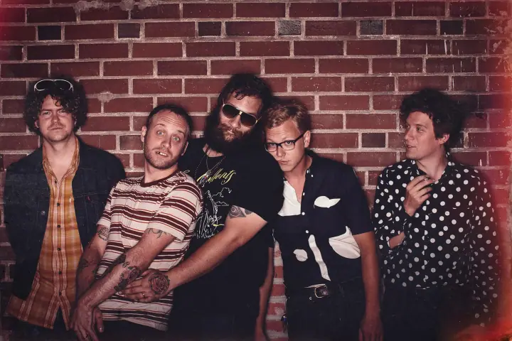 Five people pose side by side in front of a red brick wall, some wearing sunglasses and glasses.