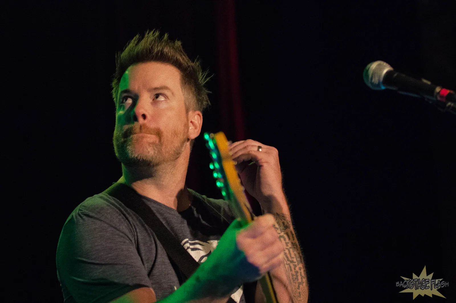 David Cook performs on stage with a guitar and a microphone at right, lit by green light against a dark backdrop.