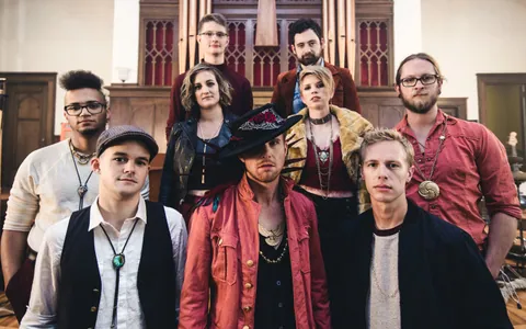 Nine people pose in a church with a pipe organ and stained glass, many in vests, hats, and layered necklaces.