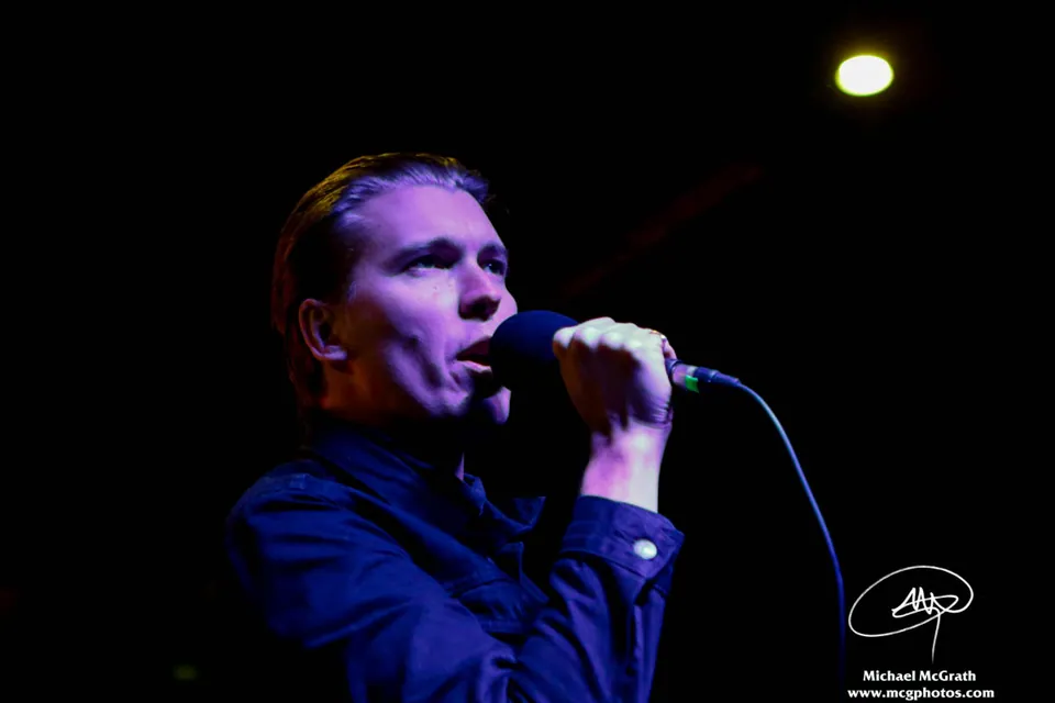 Alex Cameron sings into a microphone in a dark jacket under blue stage lighting at Globe Hall.