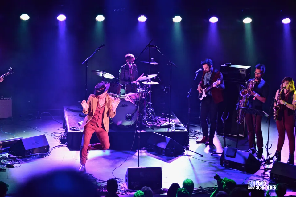 The Burroughs perform with a hatted frontman, drummer, guitarist, and horn players under purple stage lights.
