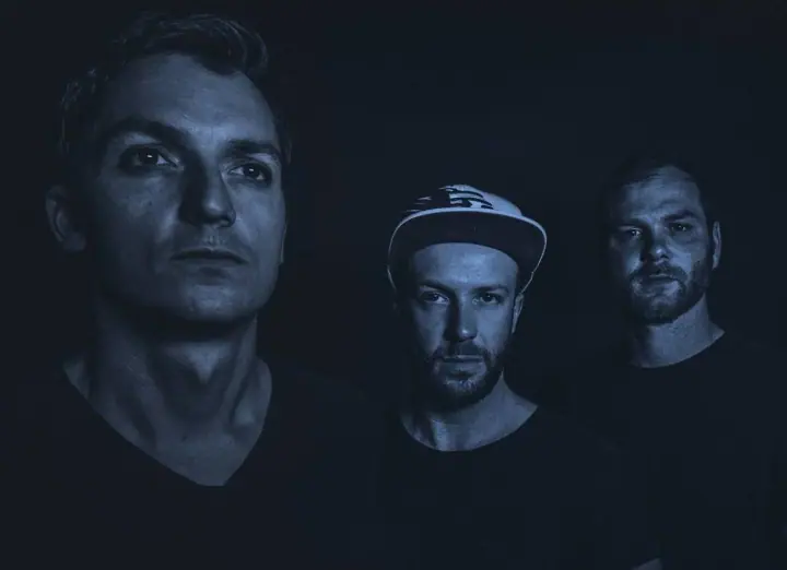 Three men in dark shirts pose against a black backdrop in a blue-toned portrait, one wearing a white cap.