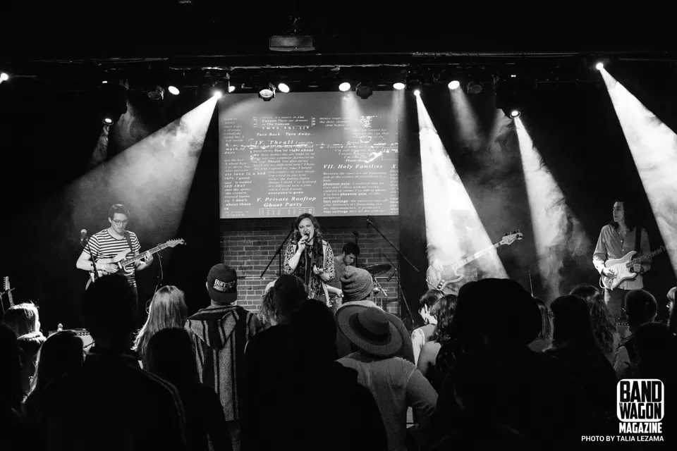 Quentin performs under spotlights with a singer, two guitarists, and a drummer, with silhouetted fans in the foreground.