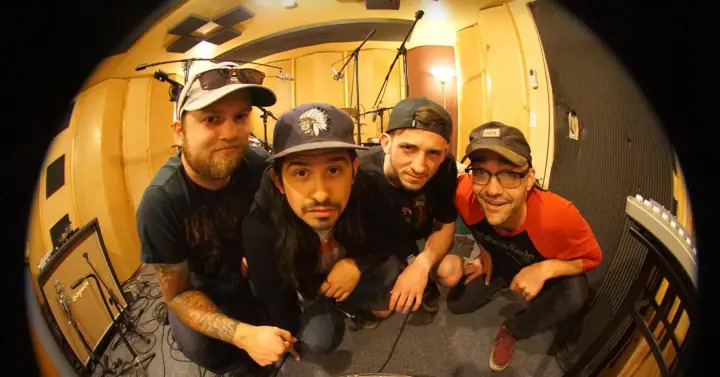 Four men crouch close together for a fisheye photo in a studio with a drum kit, mic stands, and an amplifier.