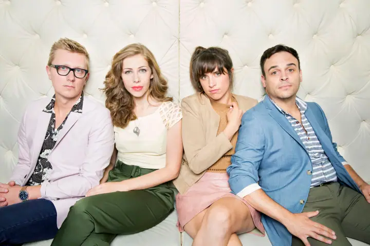 Four people in stylish casual clothing sit close together on a white tufted couch for a portrait.