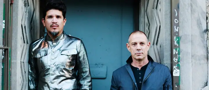 Two men pose before a teal door: one in a shiny silver jacket, the other in a navy jacket over black.