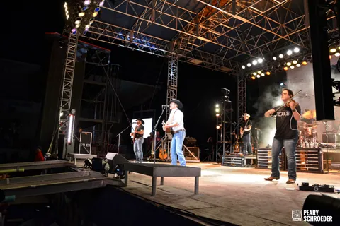 Man in a cowboy hat sings with a guitar on an outdoor stage while a fiddler in a Texas shirt plays.