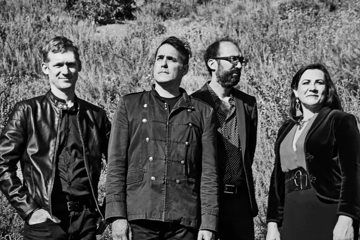 Black-and-white outdoor group portrait of four people standing in tall grass, dressed in leather and dark jackets.
