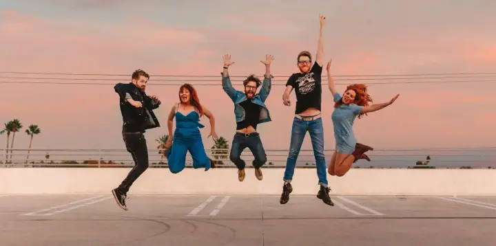 Five people leap in the air on a rooftop parking deck at sunset, with palm trees and the ocean behind them.