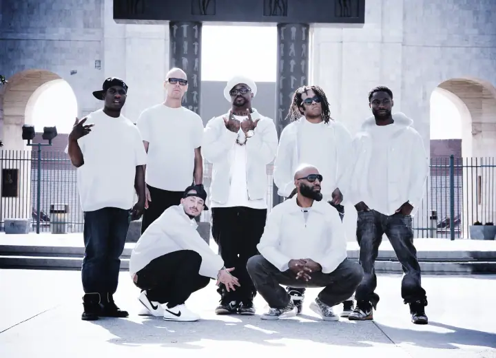 Seven men in white clothing pose outdoors in front of a stone archway and iron gate on a sunny day.