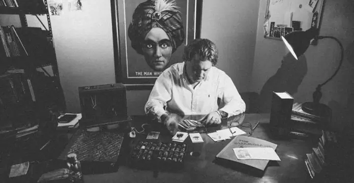 Black-and-white photo of a man in a white shirt sorting cards at a desk beneath a turbaned man's poster.