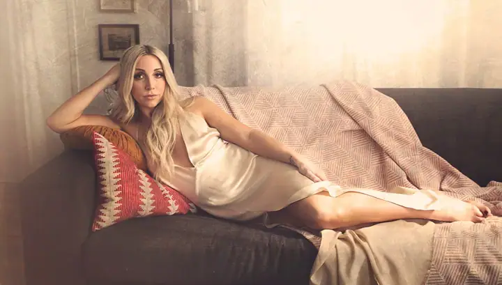 A blonde woman in a cream satin slip dress reclines on a gray sofa under a patterned throw.