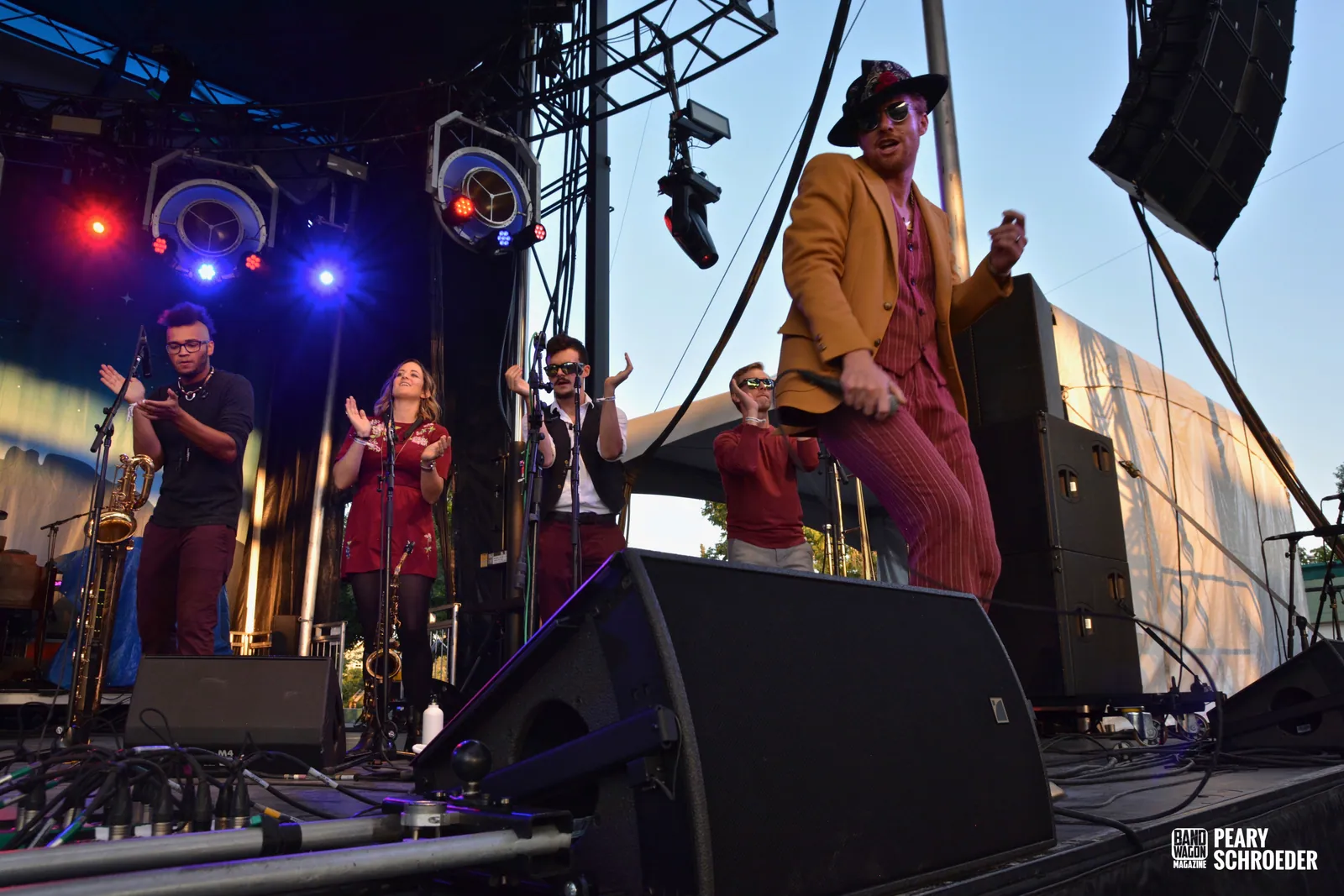 Performers in red and mustard outfits play saxophone and sing on an outdoor stage at dusk.