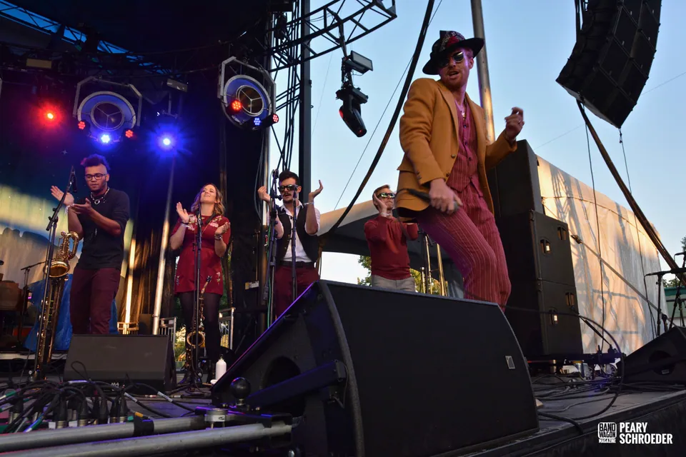 Performers in red and mustard outfits play saxophone and sing on an outdoor stage at dusk.