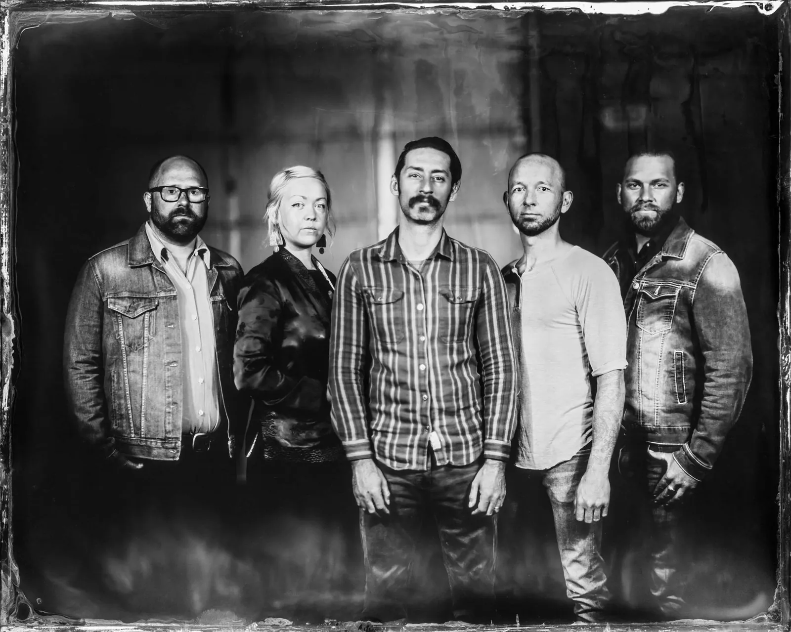 Black-and-white tintype-style group portrait of five people in denim and flannel standing against a dark backdrop.