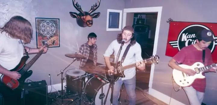 Four musicians play in a small room with a drum kit, a bass, and two electric guitars, under a mounted deer head.