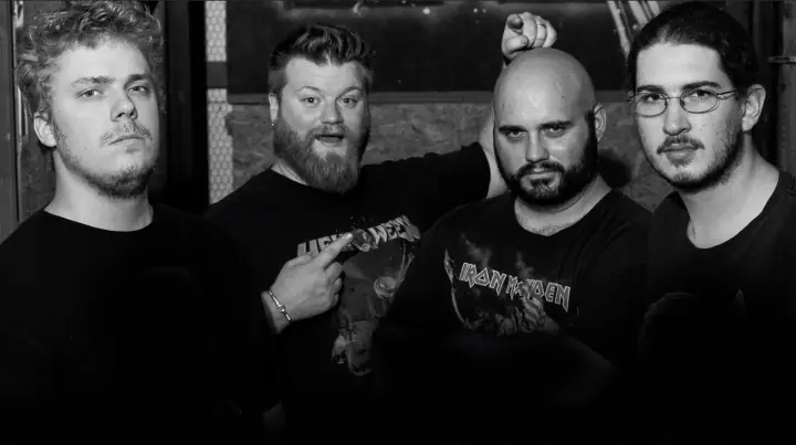 Black and white group portrait of four men in dark t-shirts posing indoors, one wearing an Iron Maiden shirt.