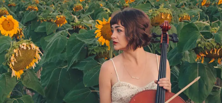A woman with a short brown bob and red lipstick holds a cello and bow in a field of sunflowers.