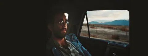 A bearded man in glasses and a denim shirt looks out a car window at a desert landscape with mountains beyond.