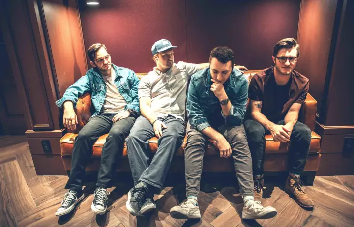 Four men sit on a brown leather couch in a wood-paneled studio, wearing denim jackets, a cap, and casual clothes.