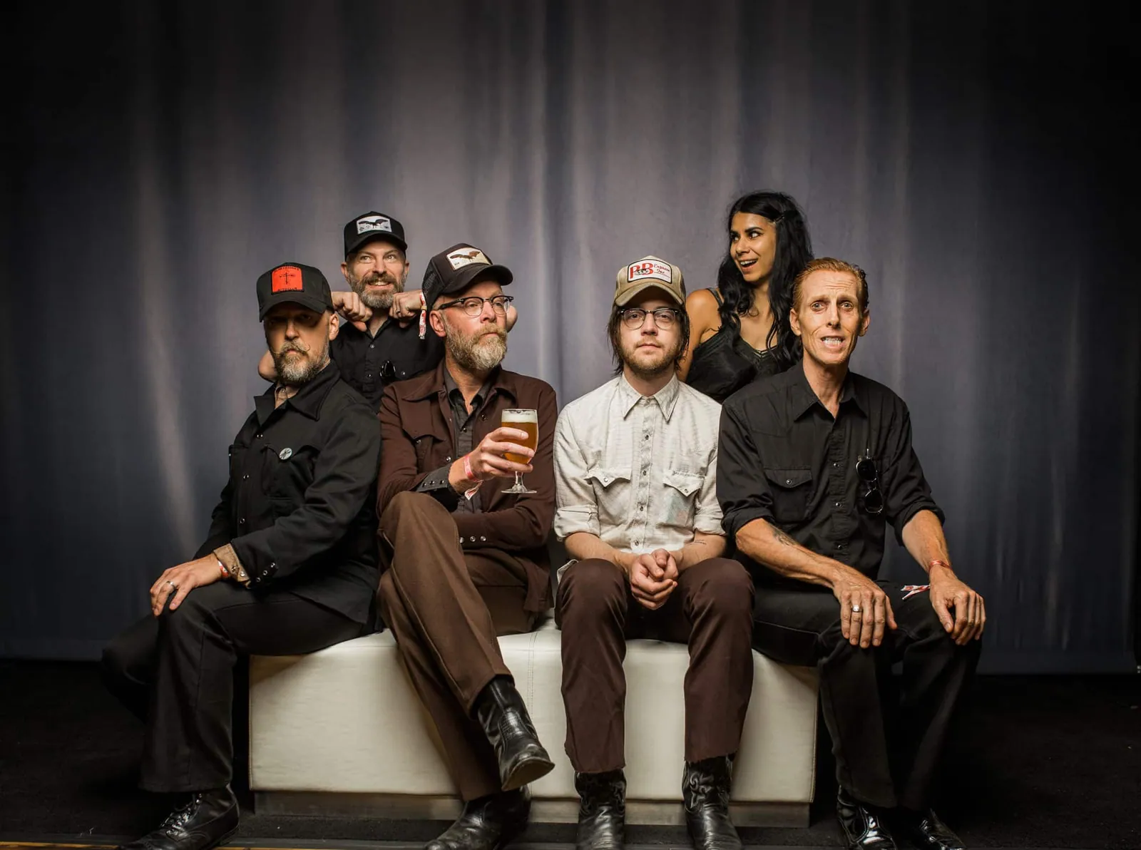 Six people in caps and dark shirts pose on a white bench before a gray curtain, one holding a beer.
