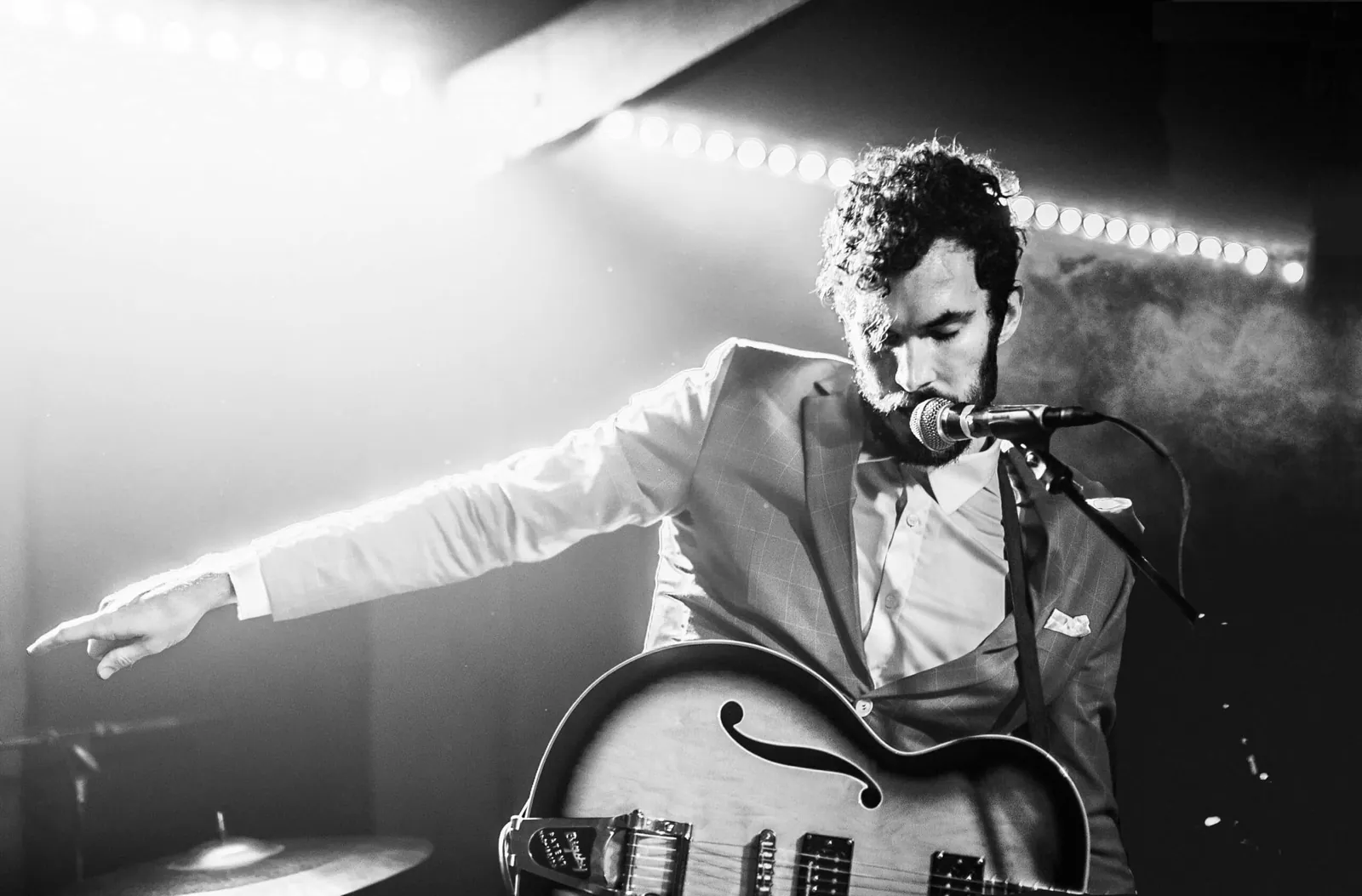 Black and white photo of a bearded singer in a suit playing a hollow-body guitar under stage lights.