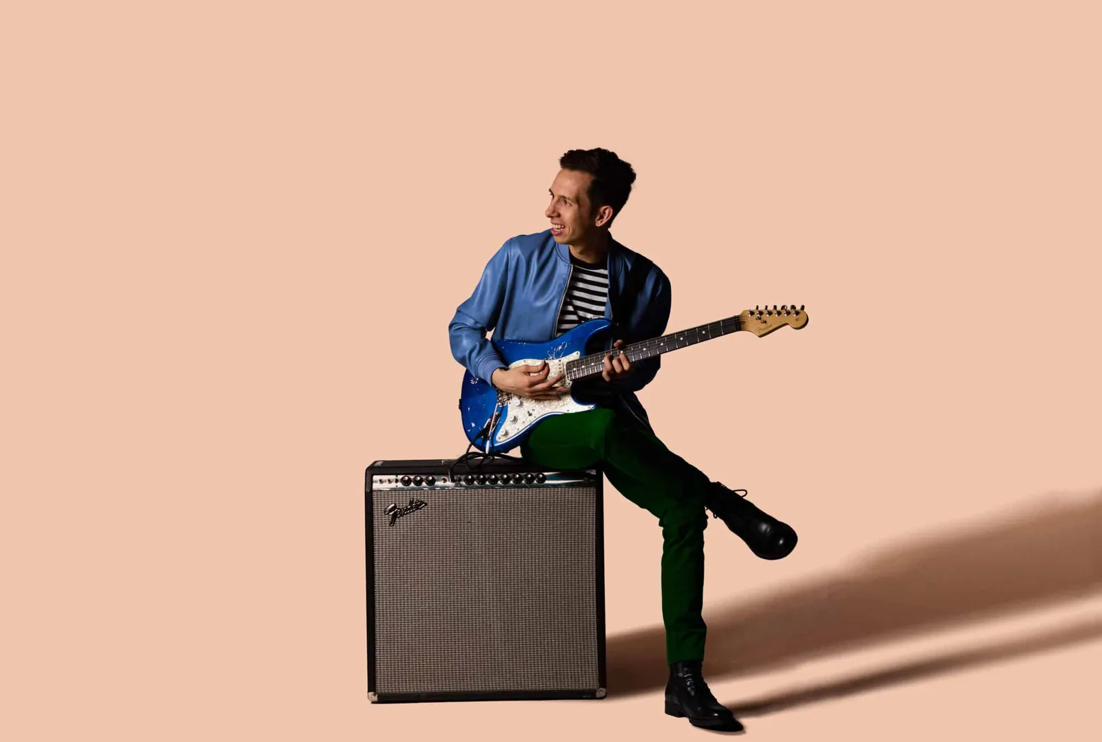 A man in a blue jacket plays a blue electric guitar while seated on a Fender amp against a peach backdrop.