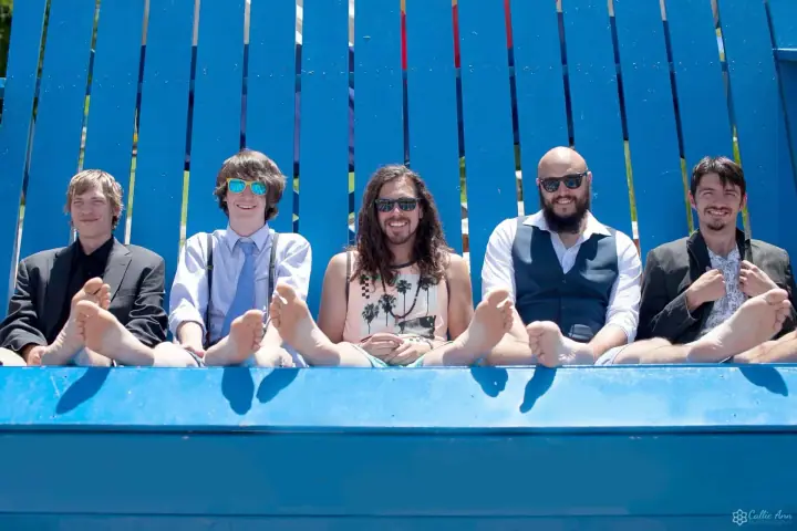 Five people sit barefoot in a row on a bright blue bench in front of a blue slatted wall on a sunny day.