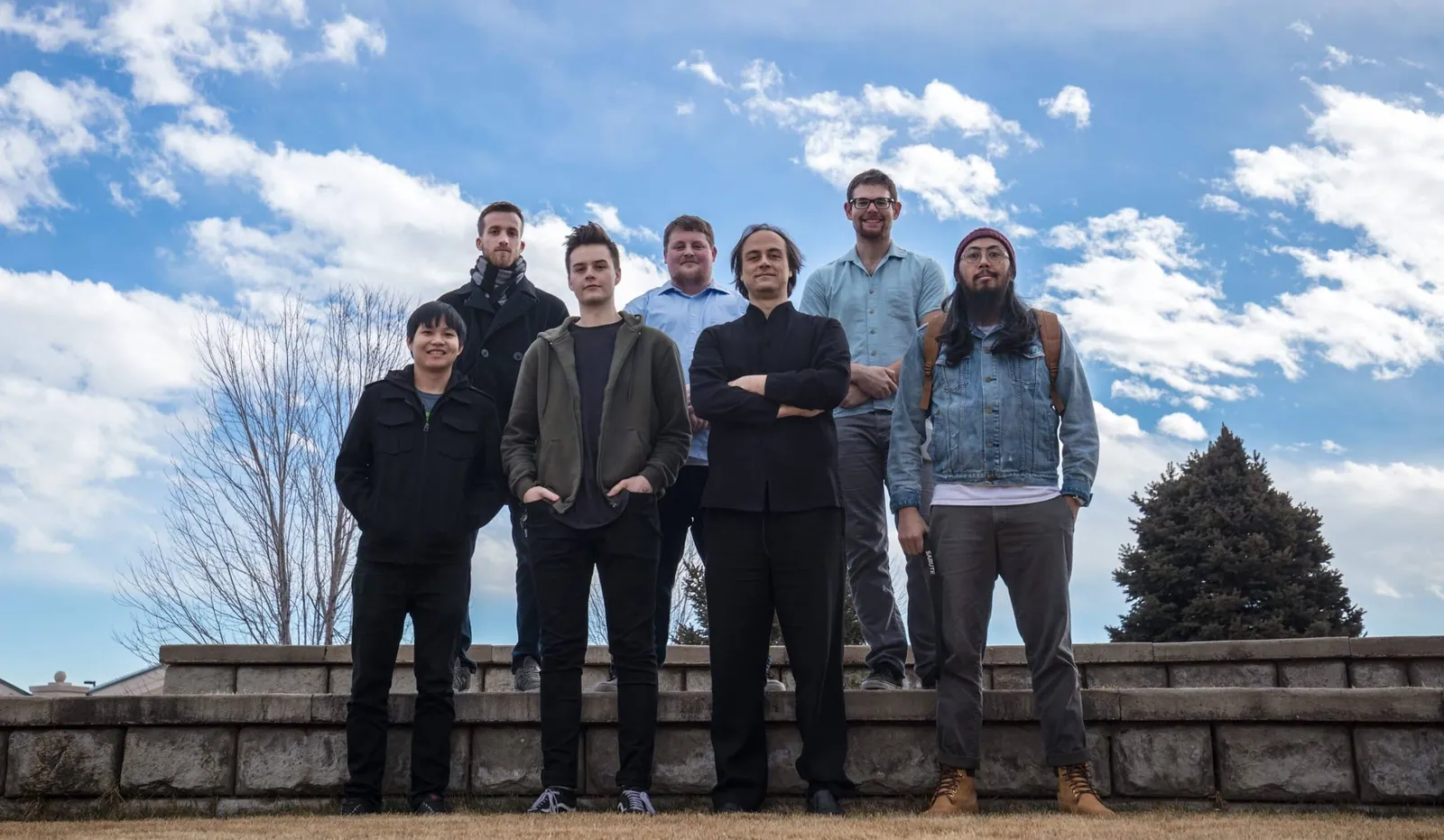 Boundless Septet poses outdoors before a stone wall and blue sky, seven members in casual dark and denim clothes.