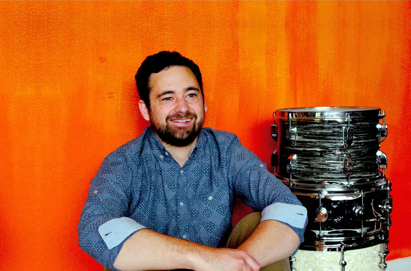 A smiling bearded man in a blue patterned shirt sits before an orange wall beside a black-and-white swirl drum kit.