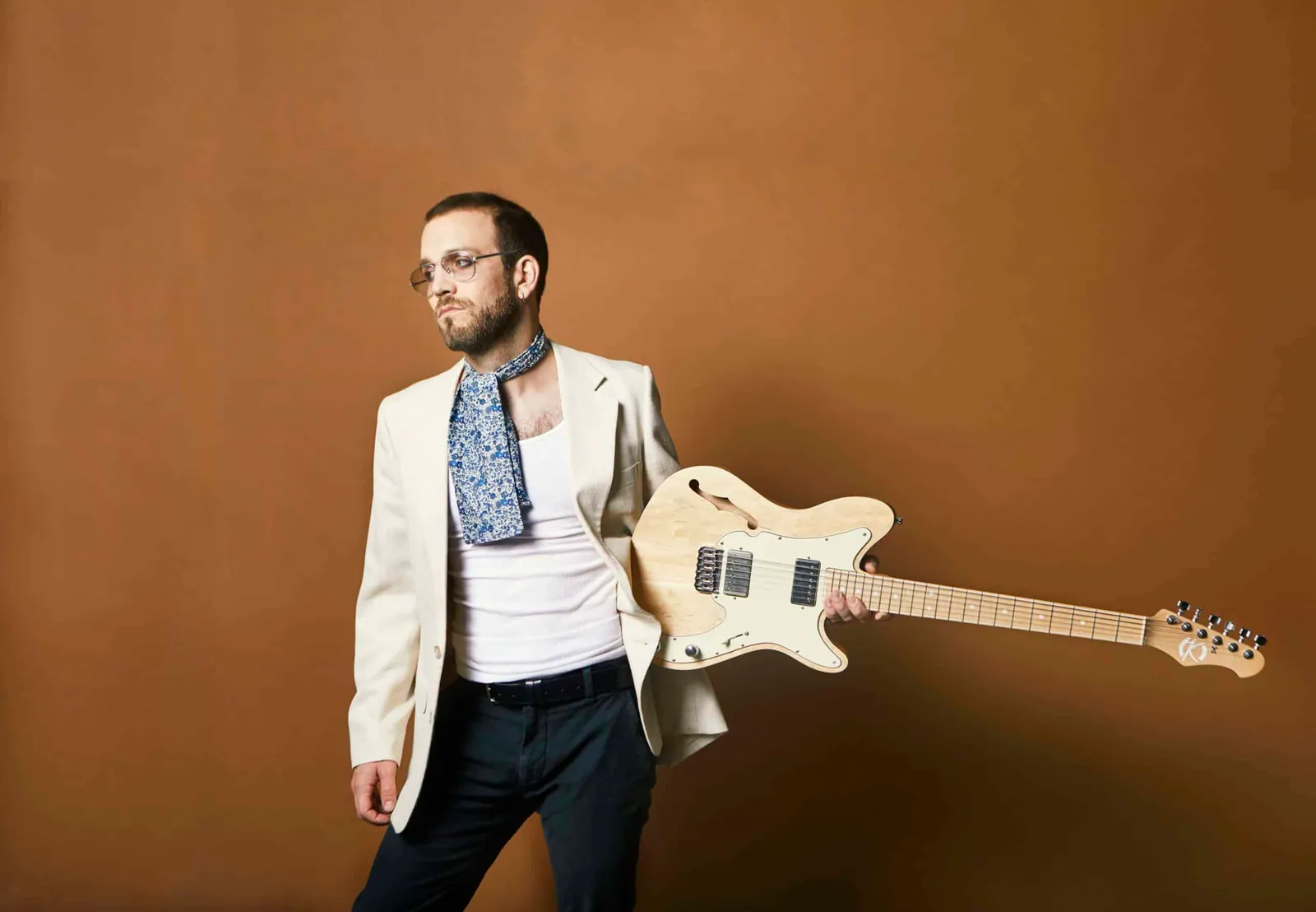 Theo Katzman in cream blazer, paisley neckerchief and glasses holds a blond electric guitar against brown backdrop.