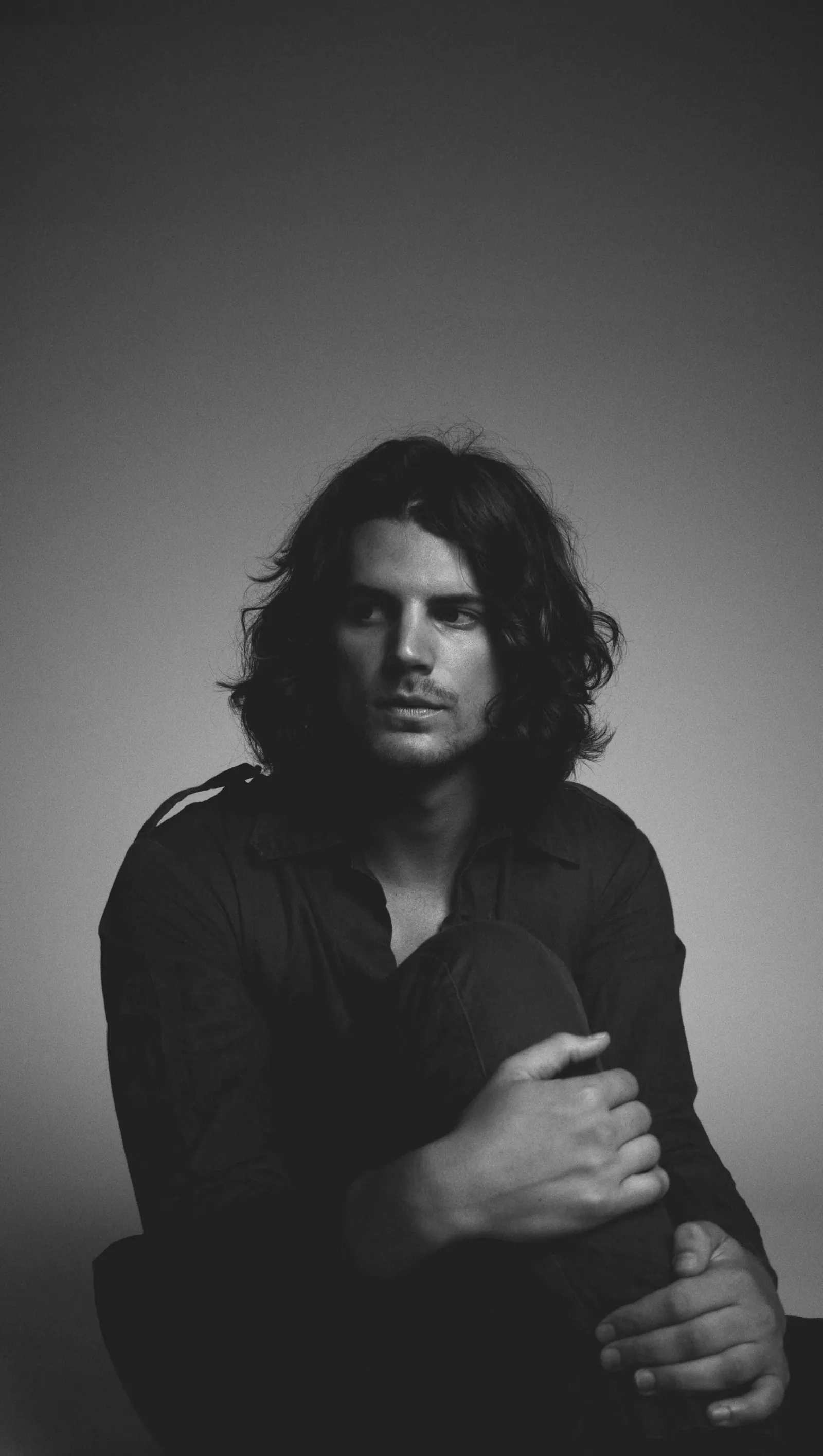 Black-and-white portrait of a long-haired man in a dark shirt, arms wrapped around his knee against a gray backdrop.