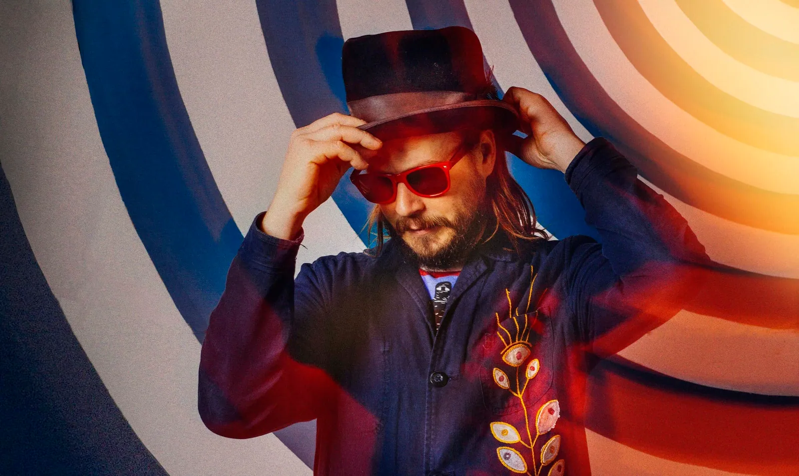 A bearded man in a dark hat and red sunglasses adjusts his hat against a bold concentric-circle backdrop.