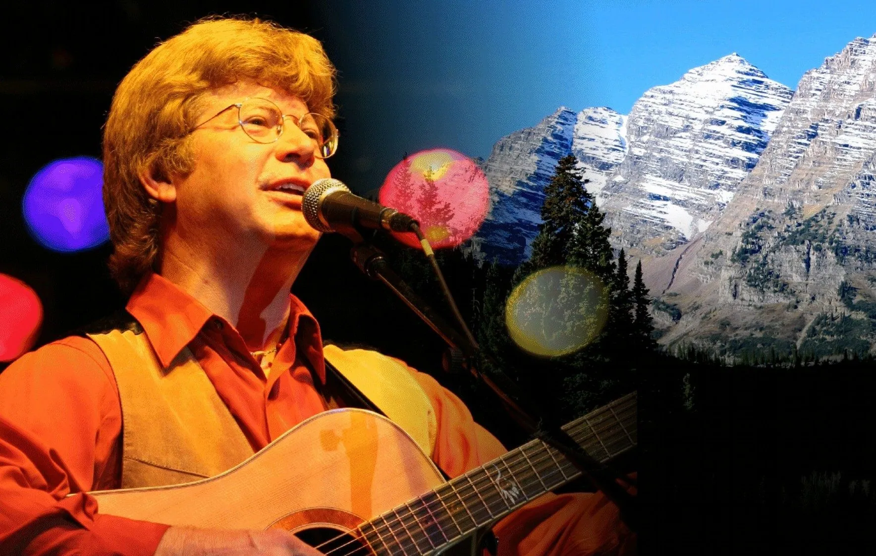 Man in glasses and a red shirt sings and plays acoustic guitar before a snowy mountain backdrop.