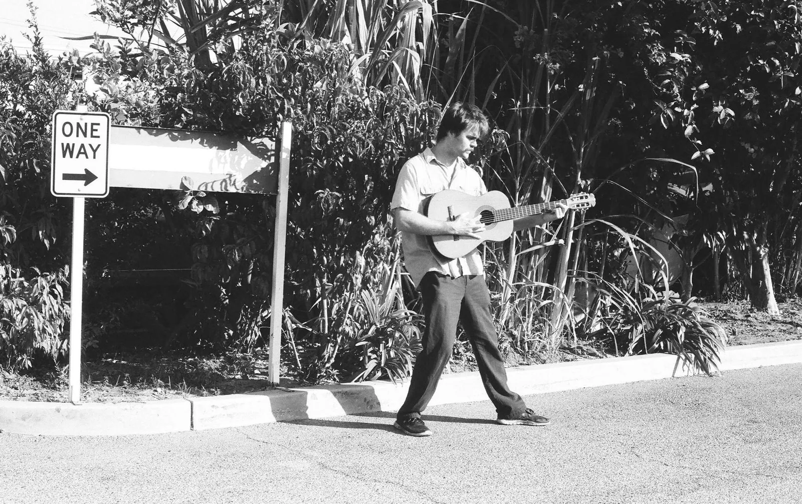 Black-and-white photo of a man in a light shirt playing acoustic guitar while walking past a One Way sign.
