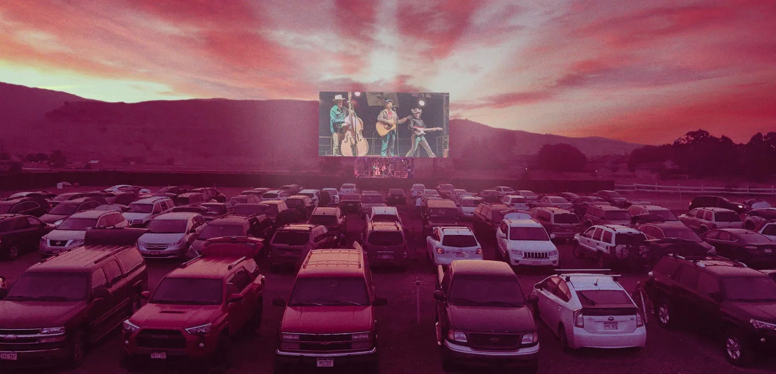 Cars fill a drive-in lot at dusk while a large screen shows three musicians playing upright bass and guitars.