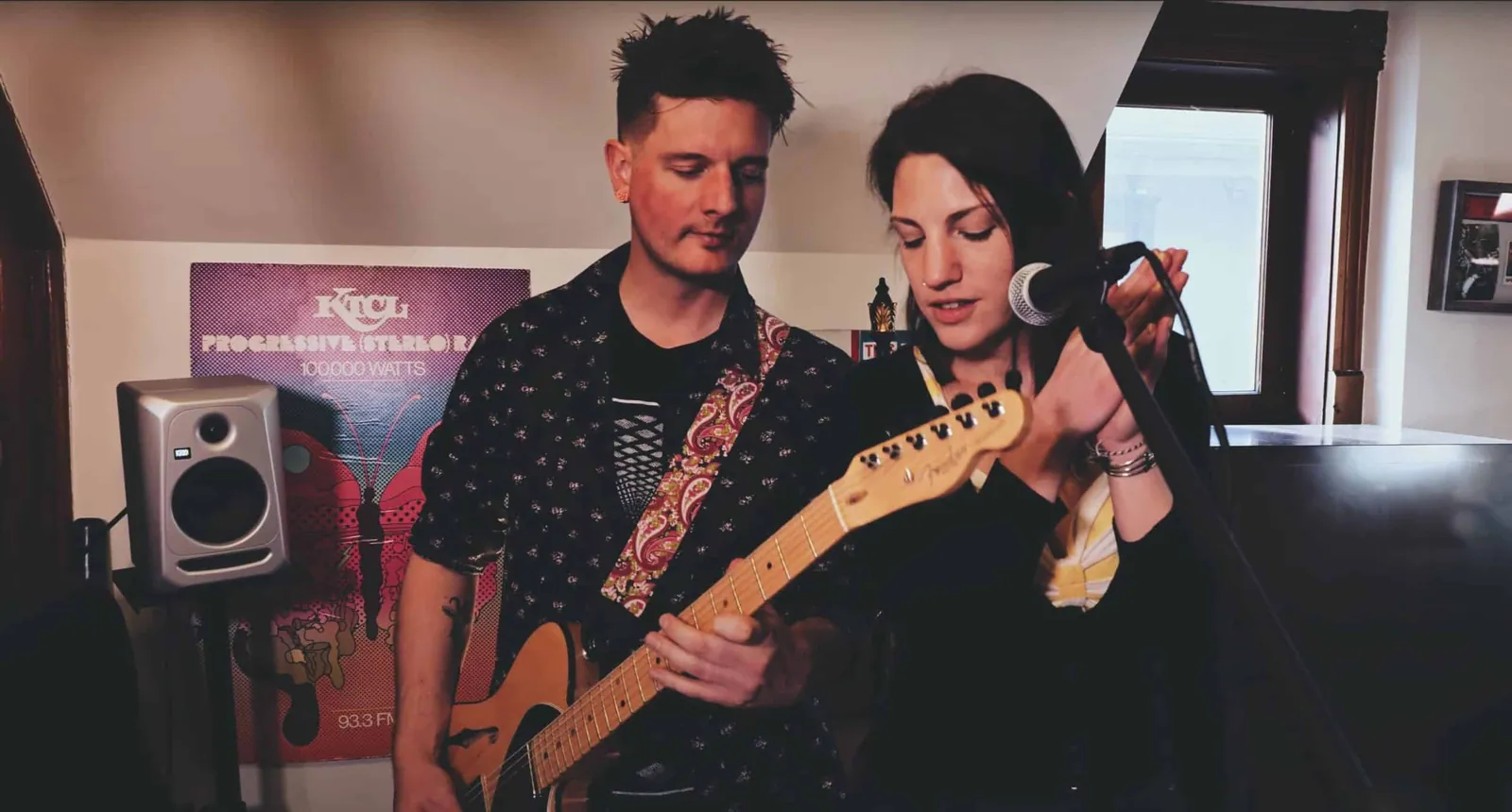 A woman sings into a microphone while a man plays an electric guitar with a paisley strap in a small studio room.