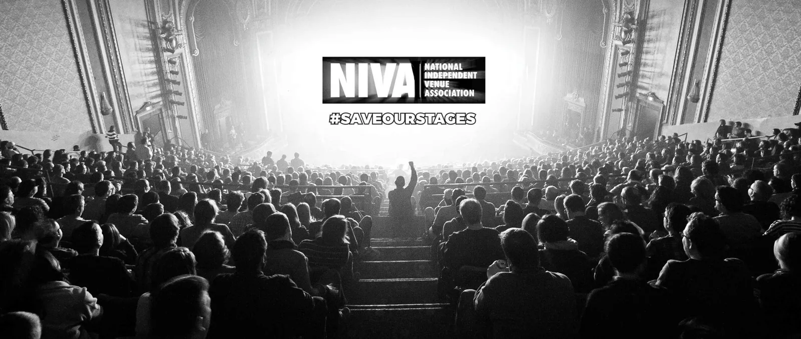 Black-and-white photo of a crowd in an ornate theater, with one person raising an arm toward a bright stage.