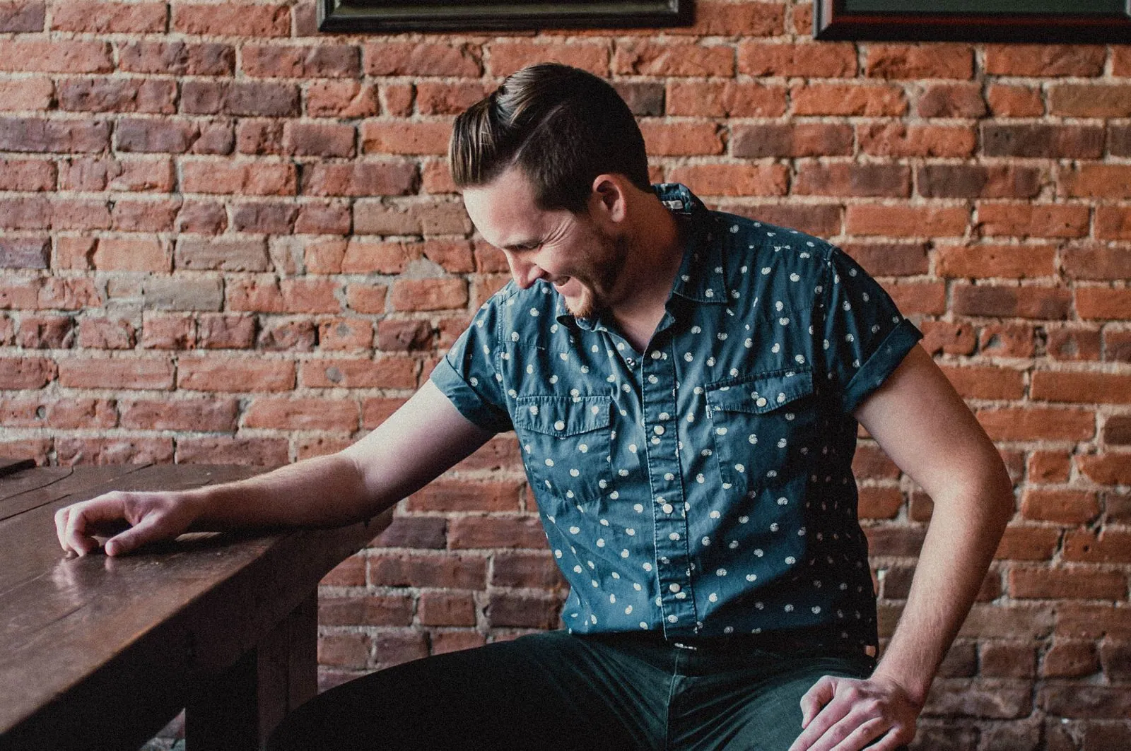 Man with swept-back hair in a teal polka-dot shirt leans on a wooden table in front of a brick wall.