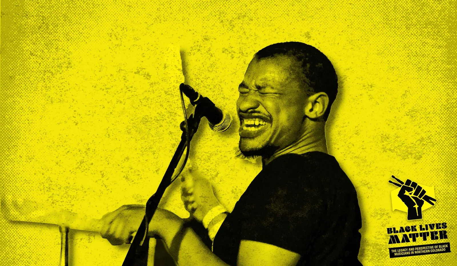 Man in a black t-shirt singing into a mic against a yellow background, with a drum kit at lower left.
