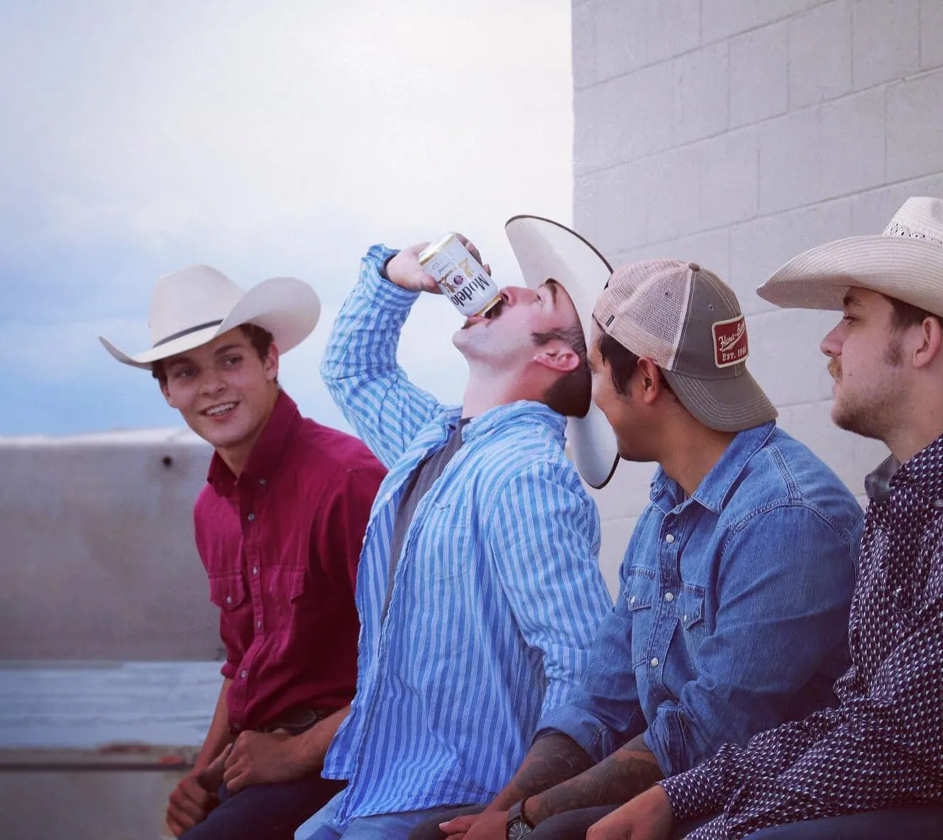 Four men in cowboy hats and casual button-up shirts sit along a concrete ledge outdoors, one drinking from a can.