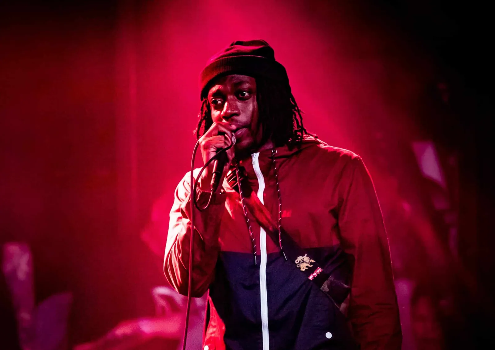 A performer in a dark beanie and zip-up jacket sings into a microphone under red stage lighting.