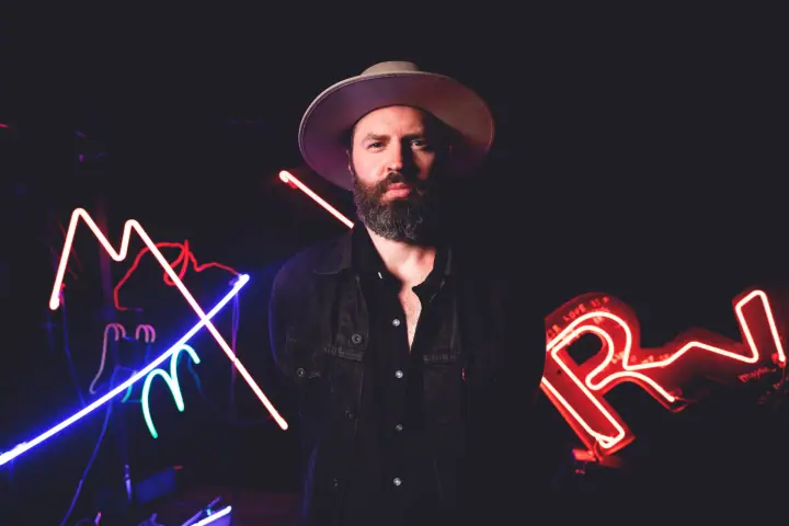 A bearded man in a wide-brim hat and dark jacket stands before glowing red and blue neon signs.