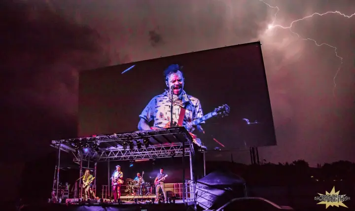 Four musicians play an outdoor stage at night under lightning as a giant screen shows a guitarist up close.