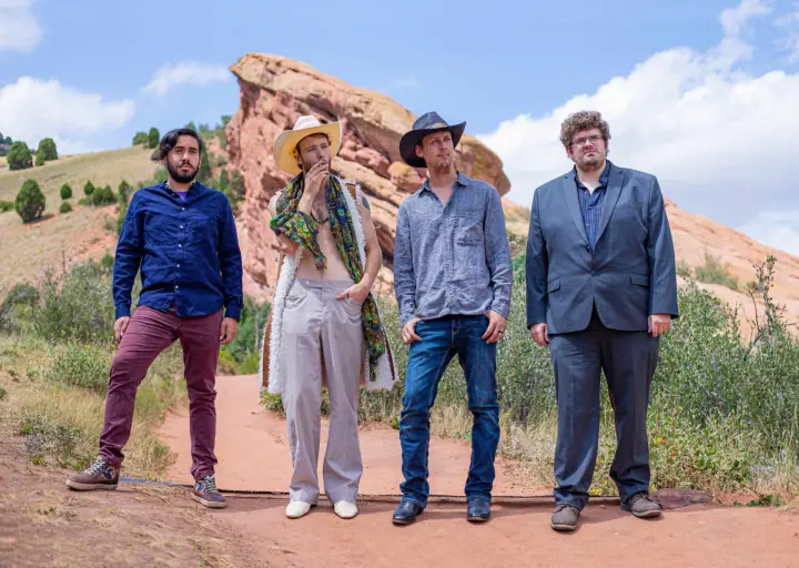 Four men pose on a dirt path in front of red sandstone rock formations, wearing hats, button-up shirts, and a blazer.
