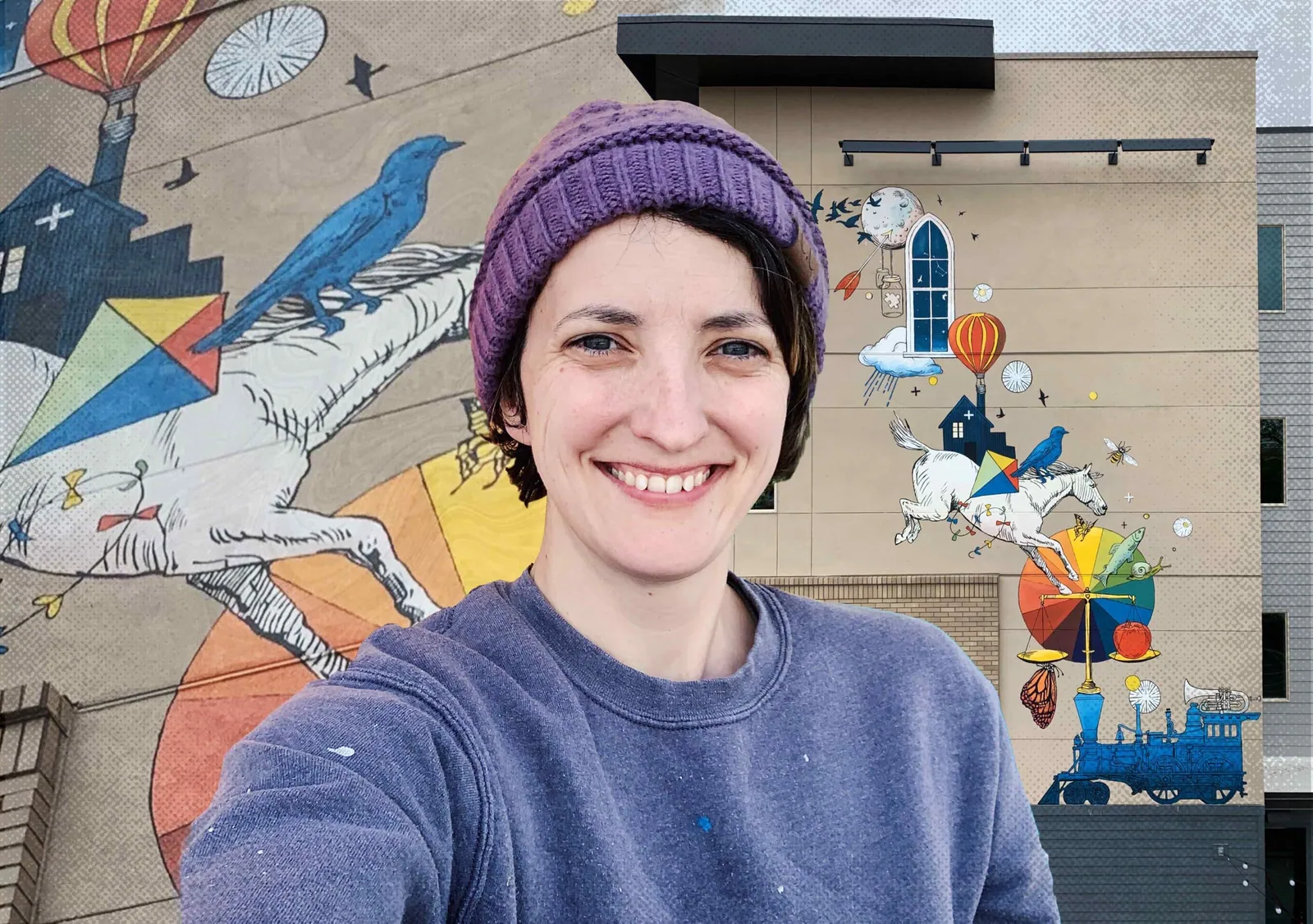 Smiling woman in a purple beanie and paint-speckled sweatshirt poses before a colorful mural with a horse and bird.