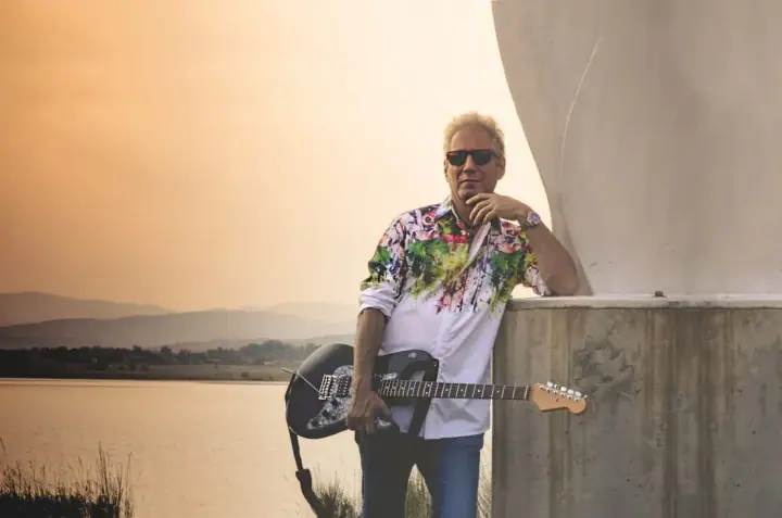 Man in sunglasses and a floral shirt holds an electric guitar and leans on a concrete wall by a lake at sunset.